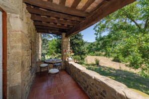 A stone-walled patio with a wooden pergola, a round table with chairs, and a view of a green landscape.