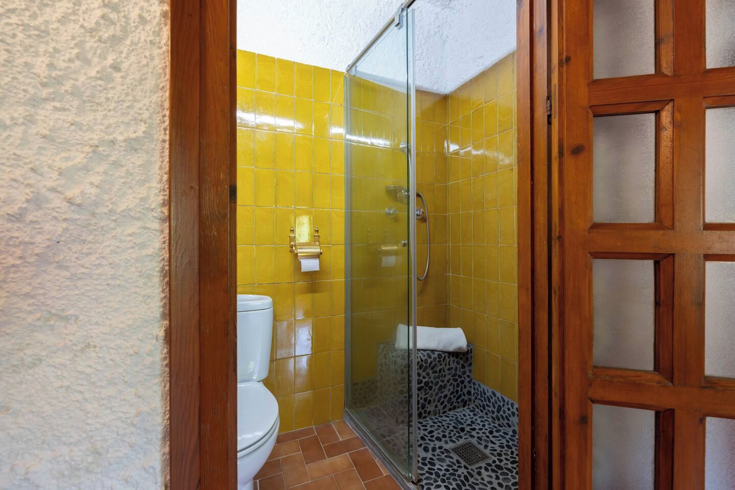 Un bagno con piastrelle gialle, cabina doccia in vetro, water bianco e una panca decorata.