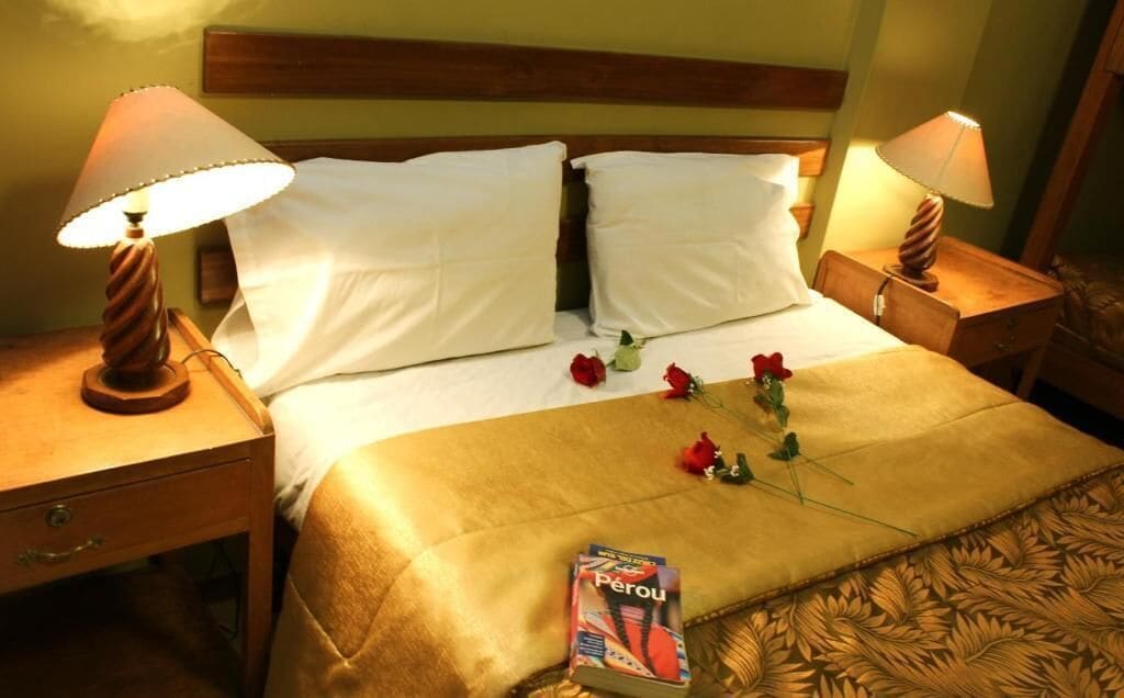 A hotel room with a bed, two bedside lamps, a nightstand, a drawer, and a book titled 'Perou' on the bed.