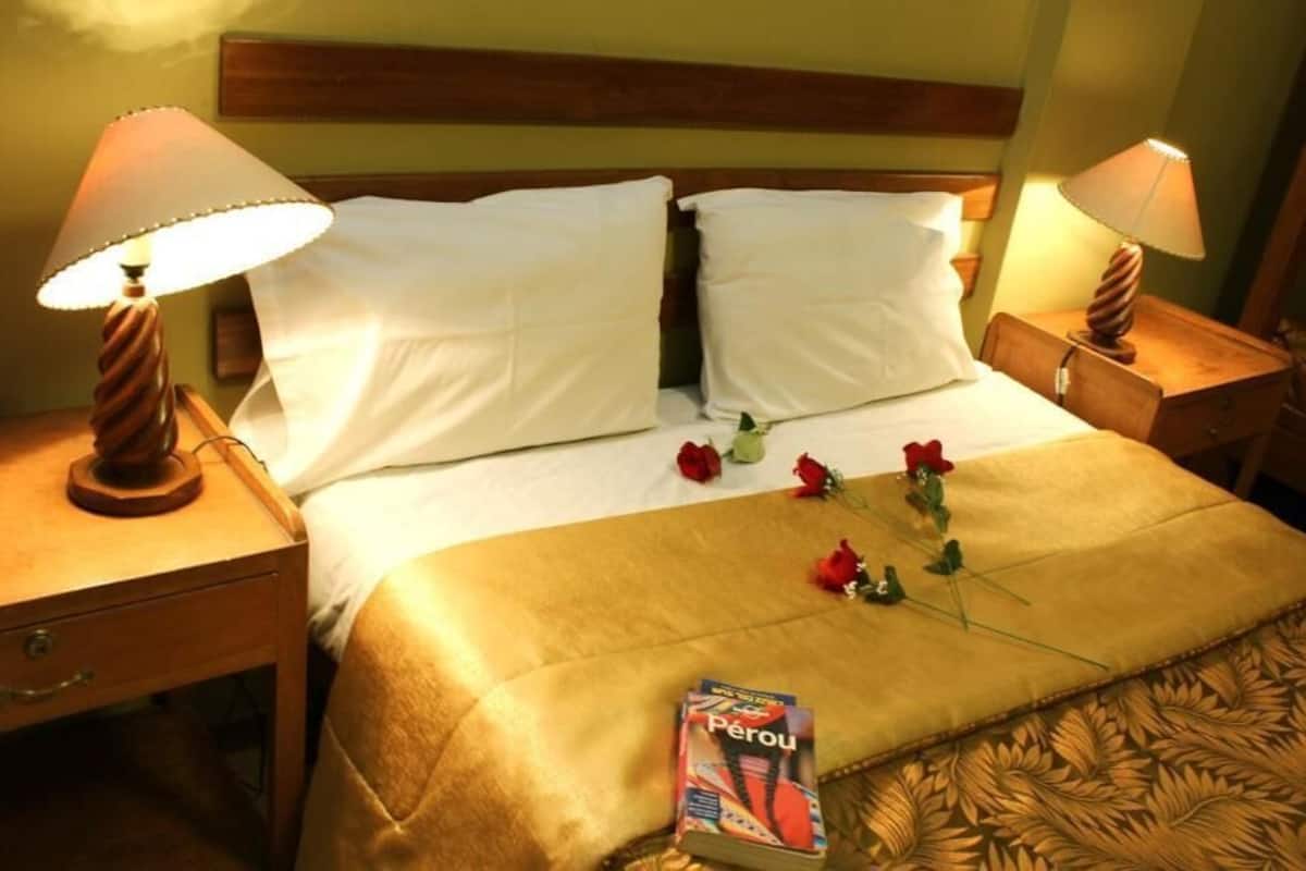 Habitación de hotel con cama, dos lámparas de noche, mesita de noche, cajón y un libro titulado 'Perou' sobre la cama.