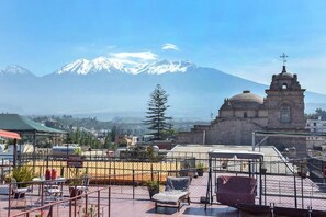 A rooftop terrace with a view of a mountain, a church, and a fenced area with chairs and tables.