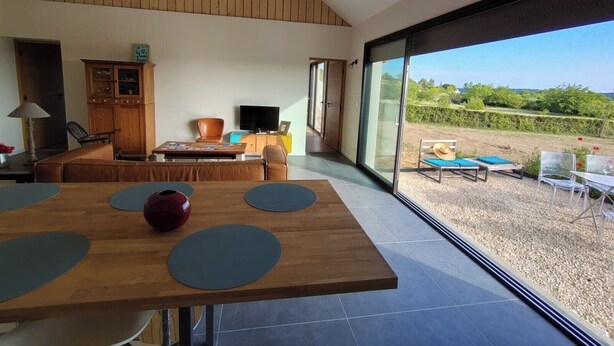 A modern living room with a wooden ceiling, a dining table with blue placemats, a television, and a view of an outdoor garden. - New house in the heart of the Loire castles - sleeps 4 (Chinon)