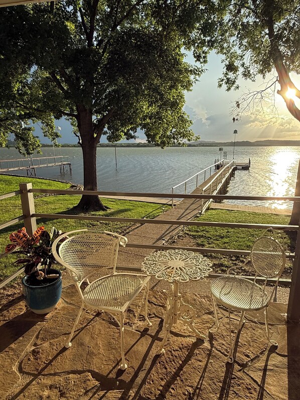 A lakeside patio with metal chairs and a table, a tree, and a dock extending into the water.