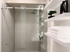 Ein modernes Badezimmer mit einer Glaskabine zur Dusche, weiß gefliesten Wänden und einem weißen Regal mit Handtüchern und einer Topfpflanze.