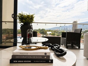 Ein modernes Zimmer mit Blick auf eine Stadtlandschaft, ein runder Tisch mit Büchern, eine Vase mit Grünpflanzen und ein dekorativer Spiegel.