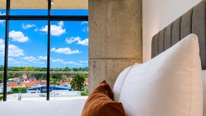 Een hotelkamer met een groot raam, een bed met witte en bruine kussens, en uitzicht op een stadsgezicht.
