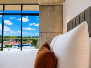 Ein Hotelzimmer mit großem Fenster, einem Bett mit weißen und braunen Kissen und Blick auf eine Stadtlandschaft.