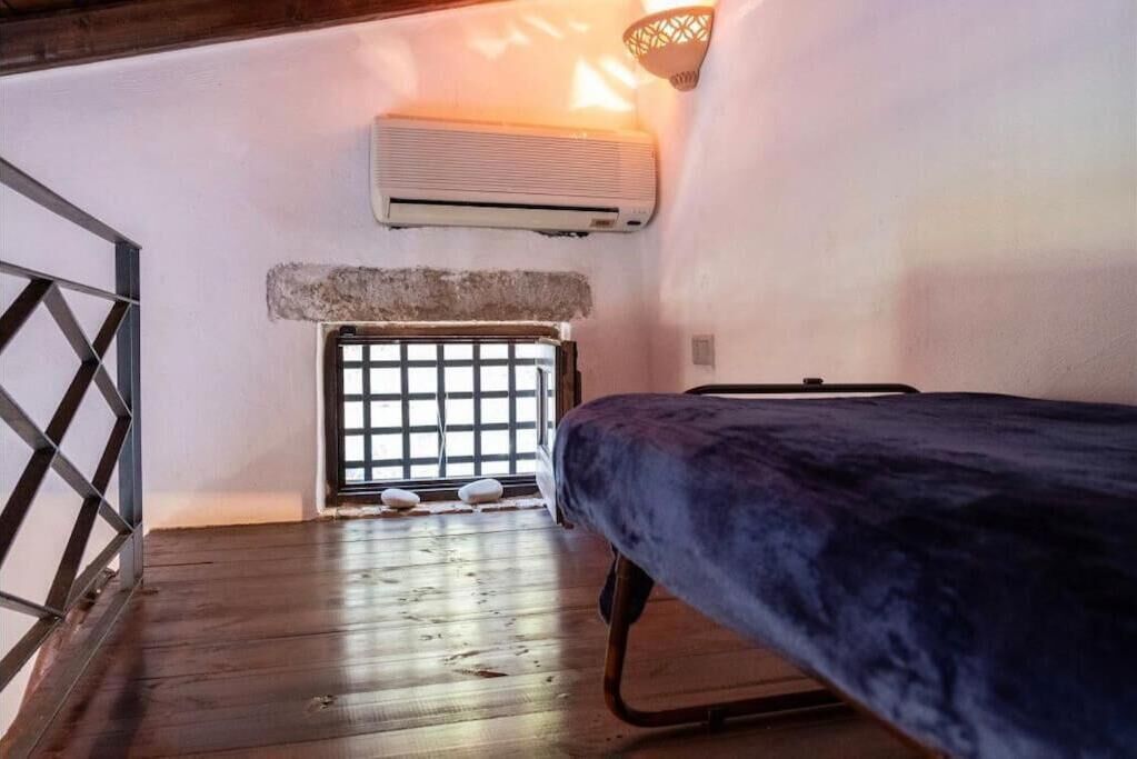 A room with a bed, an air conditioner, a window with a grid pattern, and a wooden floor.