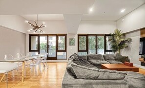 A modern living room with a sectional sofa, a dining table with chairs, and a large window with a view of greenery.