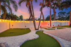 A landscaped area with artificial grass, palm trees, and a white wall with string lights.