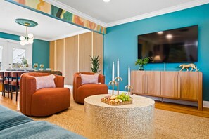 Ein Wohnzimmer mit einer türkisfarbenen Wand, einem Flachbildfernseher, einer Holzkommode und zwei orangefarbenen Sesseln.