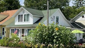 Uma casa de dois andares com telhado verde, uma bandeira e um jardim com girassóis.