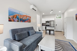 A modern living room with a grey sofa, a white coffee table, and a kitchen area with white cabinets and stainless steel appliances.