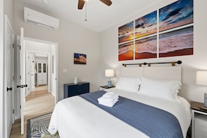 A hotel room with a bed, a nightstand, a lamp, a painting of a sunset, and an air conditioning unit.