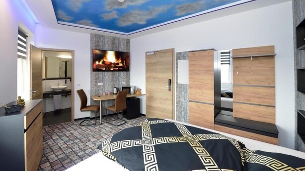 A modern bedroom with a ceiling mural, a bed with a patterned bedspread, a desk with a chair, and built-in wardrobes.