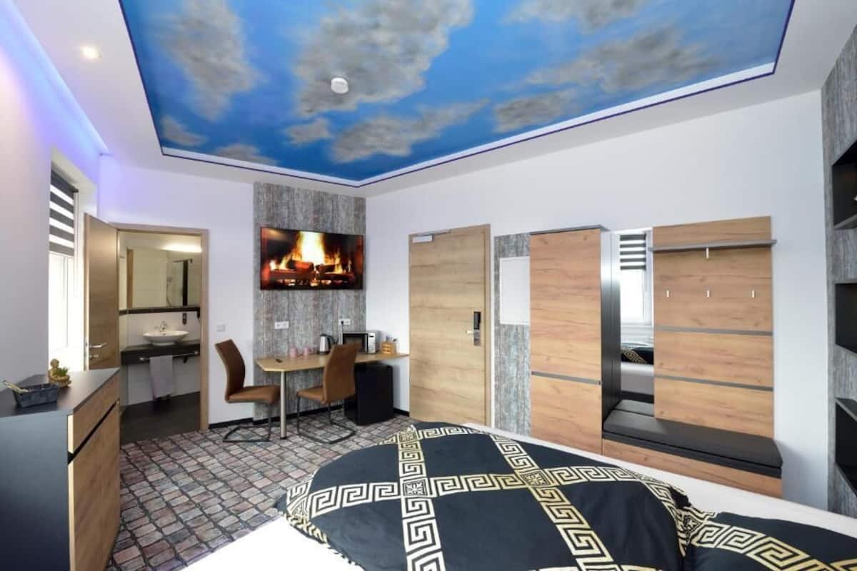 A modern bedroom with a ceiling mural, a bed with a patterned bedspread, a desk with a chair, and built-in wardrobes.