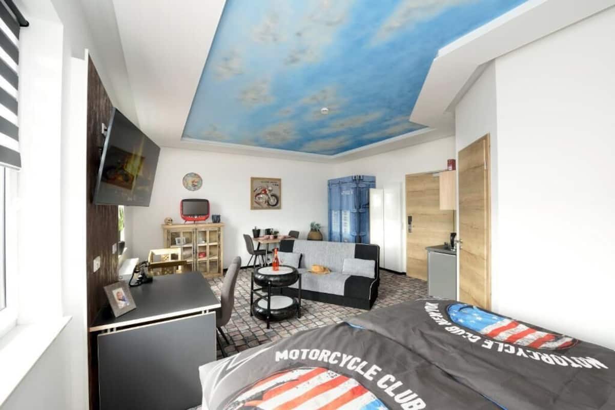 A room with a motorcycle-themed bed, a ceiling with a cloud design, a TV mounted on the wall, and a small dining area with a table and chairs.