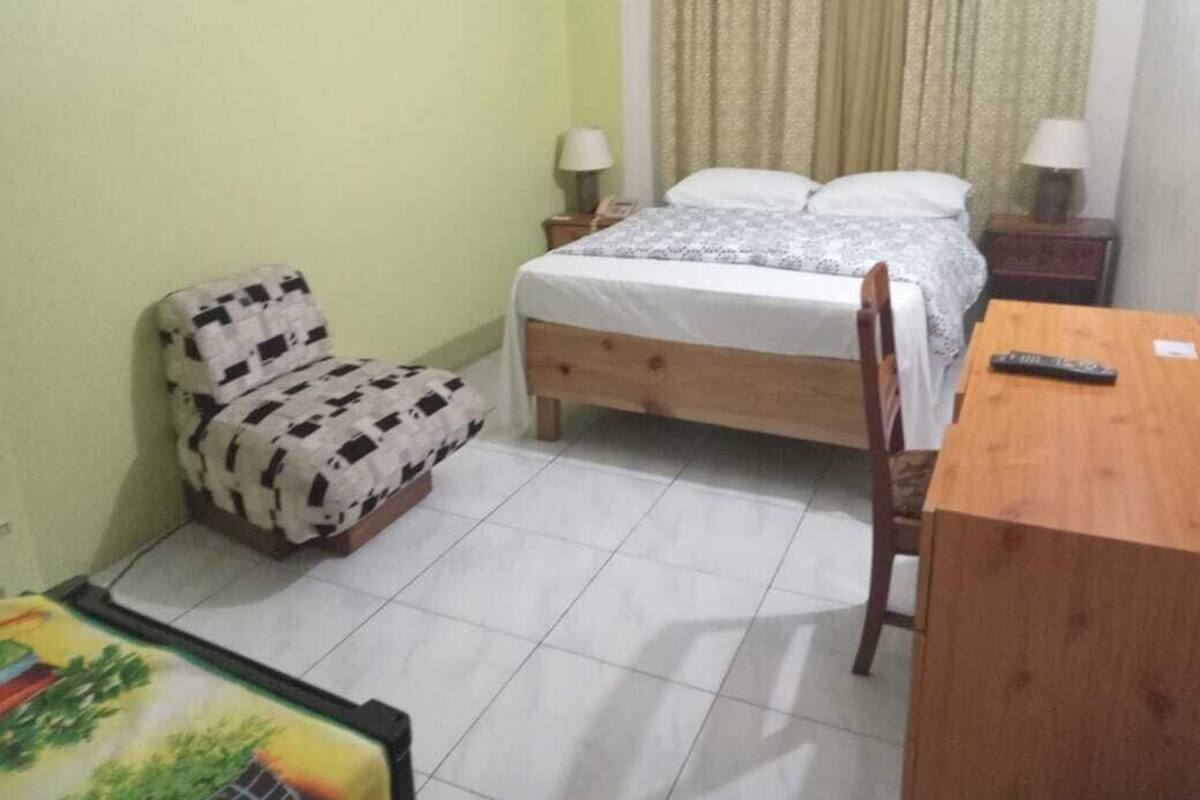 A small, clean room with a bed, a chair, a wooden table, and a nightstand with a lamp.
