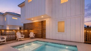 Una piscina con bordo in legno e sedie, circondata da un edificio bianco con recinzione in legno.