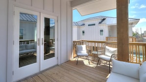 Una terrazza esterna coperta con pavimento in legno, pareti bianche e una porta vetrata che dà su un ambiente con un divano e un tavolo.