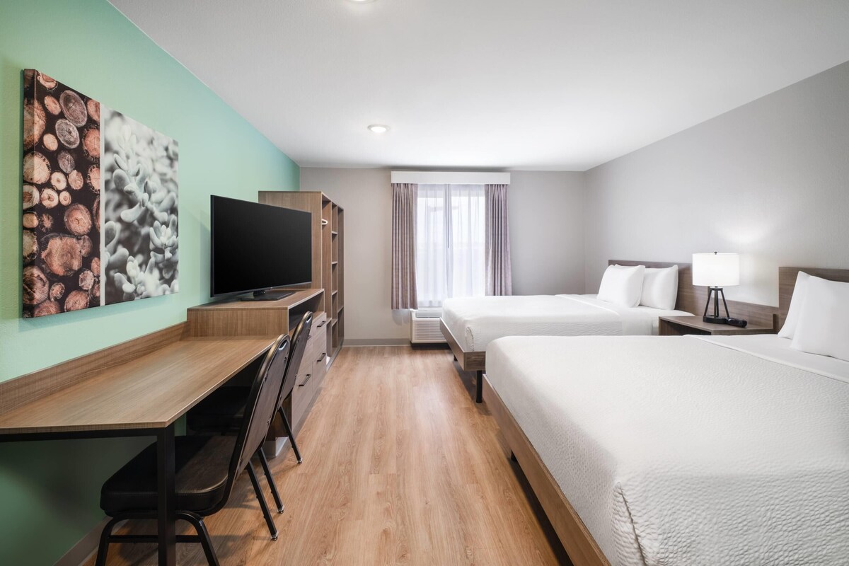 Habitación de hotel con escritorio de madera, televisor de pantalla plana, dos camas, ventana con cortinas y una obra de arte en la pared.
