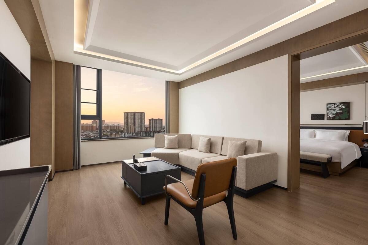 A modern hotel room with a large window, a sofa, a coffee table, a TV, and a bed.