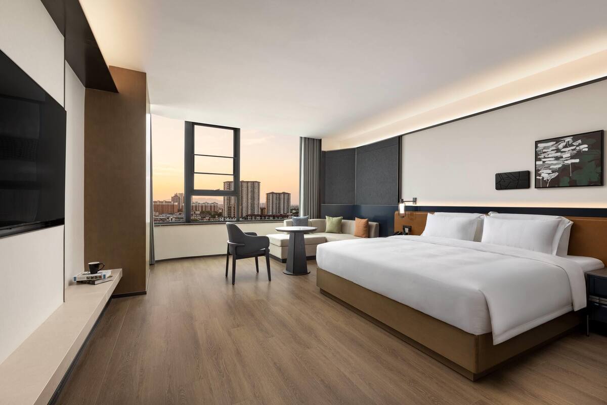 A modern hotel room with a large bed, a desk, a chair, a small table, and a city view.