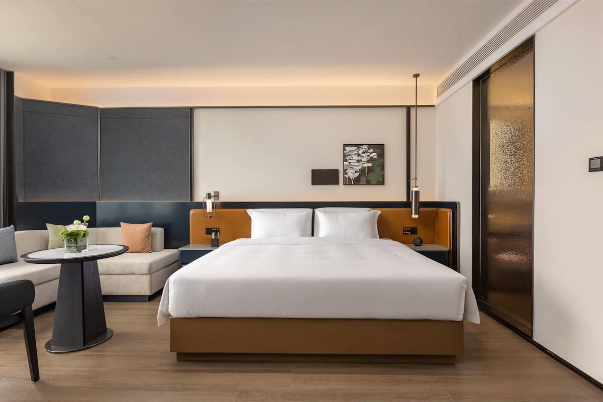 A modern hotel room with a large bed, a sofa, a small table with flowers, and a wall-mounted artwork.