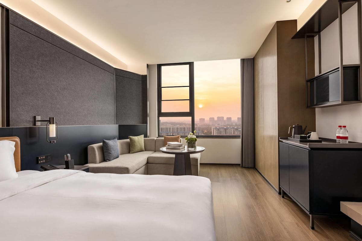 A modern hotel room with a large bed, a sofa, a small table with a vase, a city view, and a kitchenette.