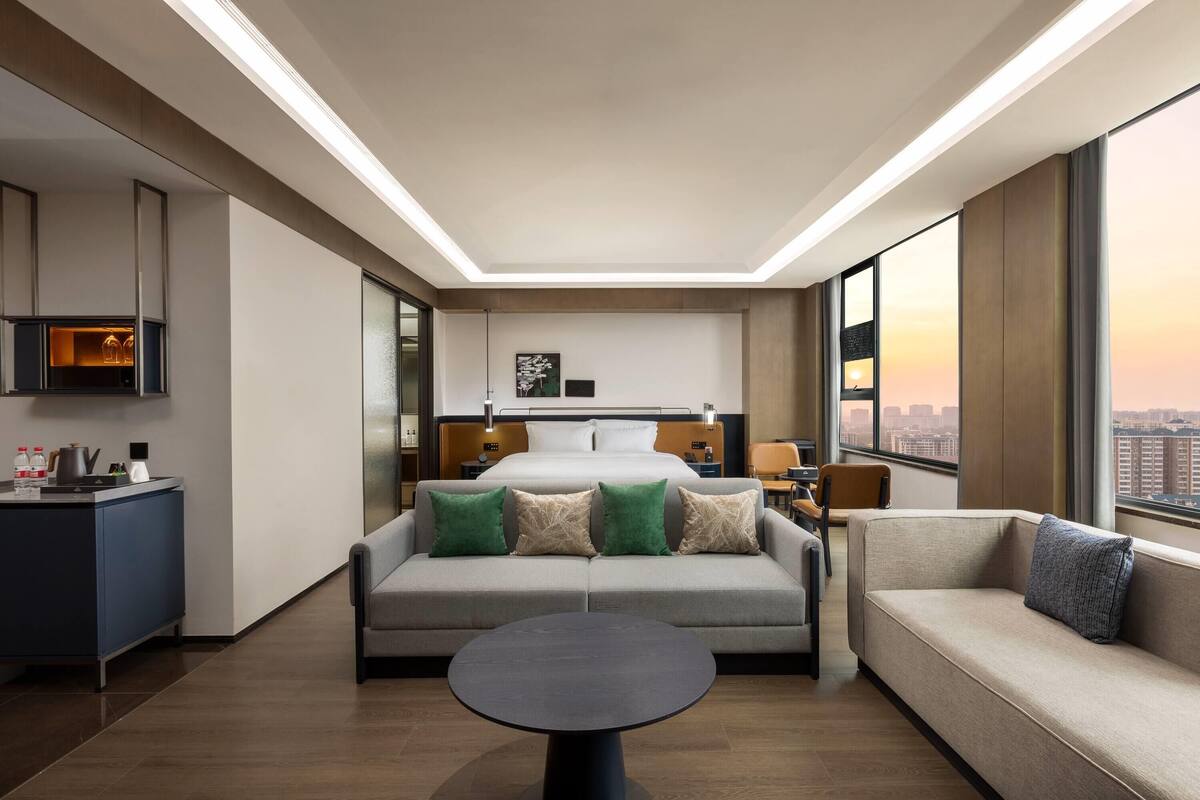 A modern hotel room with a bed, two sofas, a round table, and a city view.