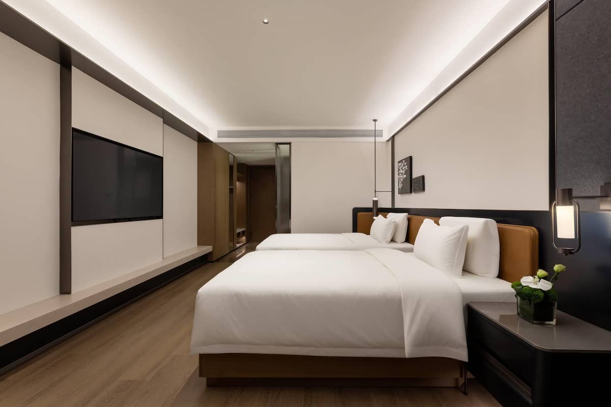 A hotel room with two beds, a flat-screen TV, a bedside table with a vase of flowers, and a wall-mounted light fixture.