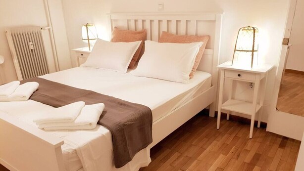Een netjes opgemaakt bed met wit en bruin beddengoed, een nachtkastje met een lamp, en een houten vloer.
