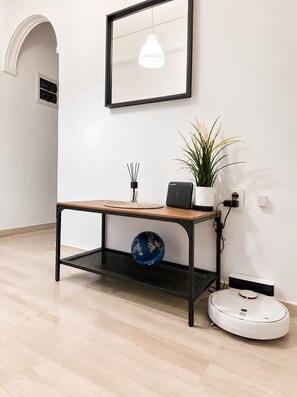 A minimalist room with a wooden table, a blue decorative ball, a white plant, a wall-mounted light fixture, and a white robotic vacuum cleaner. - Quiet Marina Zea, 3-Min Walk & Self Check-in (Piraeus)