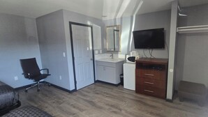 A hotel room with a wooden floor, a black office chair, a white desk with drawers, a flat-screen TV mounted above the desk, and a door leading to another room.