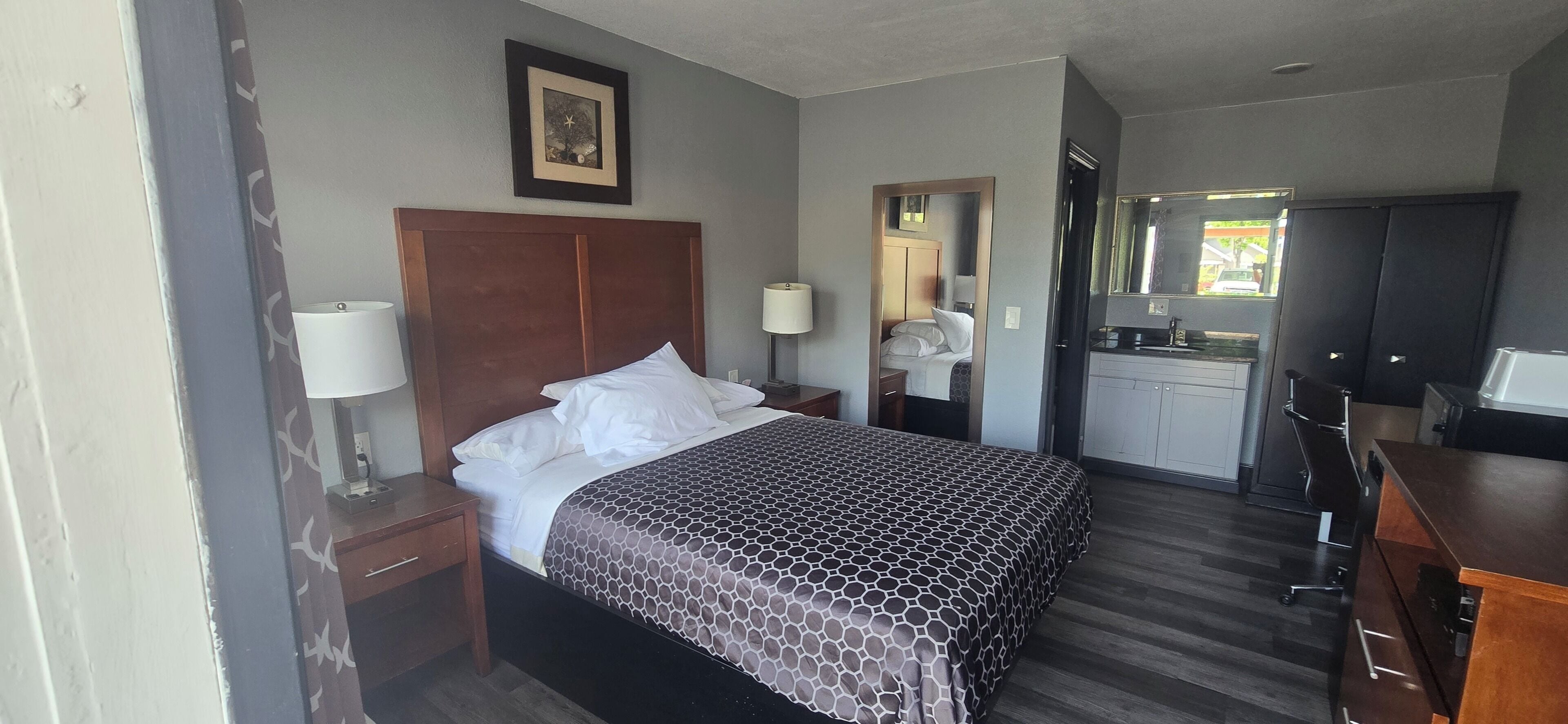 Habitación de hotel con cama, mesita de noche, escritorio, espejo, armario y kitchenette.
