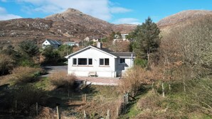 A white house with a sloped roof, surrounded by trees and a mountainous backdrop. - Woodview (Isle of Barra)