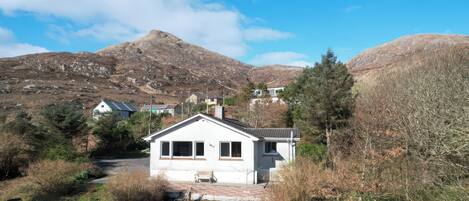 A white house with a sloped roof, surrounded by trees and a mountainous backdrop.