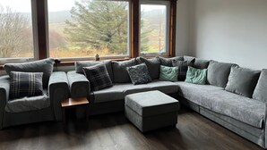 A modern living room with a sectional sofa, ottoman, and a view of a forest through large windows. - Woodview (Isle of Barra)