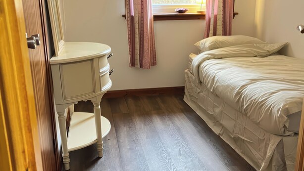 A bedroom with a bed, a bedside table, a window with curtains, and a wooden floor. - Woodview (Isle of Barra)