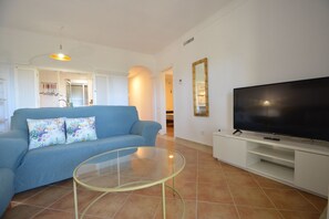 Ein Wohnzimmer mit Sofa, einem Glastisch, einem Fernseher und einer Küchenzeile im Hintergrund.