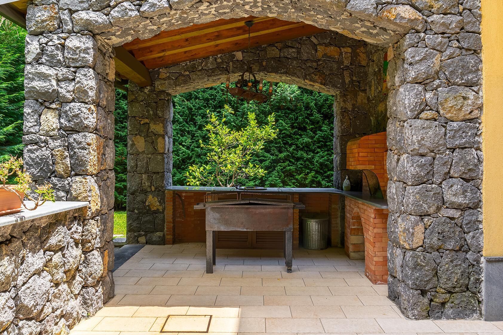 Cocina exterior de piedra con horno de ladrillo, rodeada de muros de piedra y una pérgola de madera.