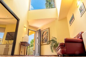 Ein Zimmer mit großem Fenster, einer zur Außenseite führenden Tür, einem Fernseher, einem Schreibtisch, einem Sessel und einem Gemälde an der Wand.