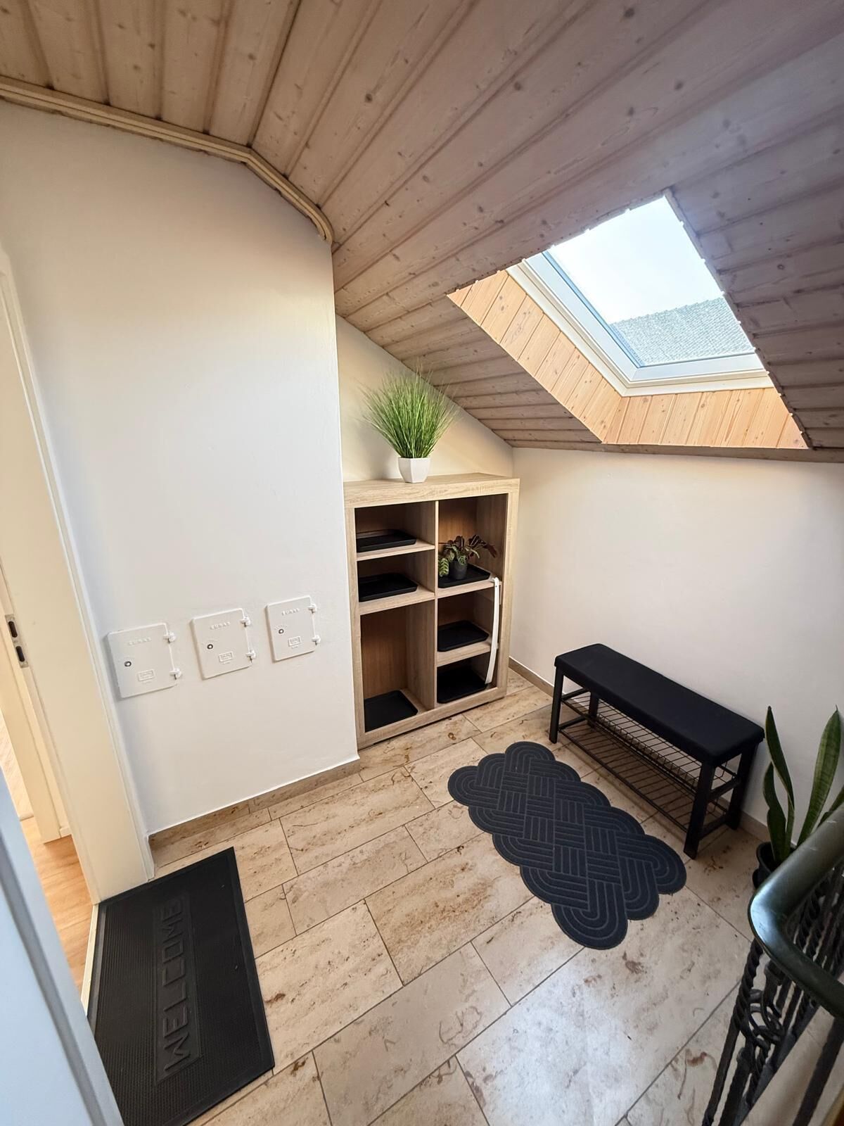 Een kamer met een dakvenster, een houten bankje, een zwart vloerkleed en een plant.