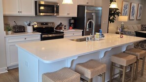 A modern kitchen with white cabinetry, stainless steel appliances, a large island with bar stools, pendant lighting, and a dining area with a sofa and wall art. - Coastal Luxury Townhome in West Ocean City, 4 Bedrooms , Near Beach, Assateague (Berlin)
