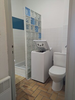 A bathroom with a white toilet, a white pedestal sink, a shower with a glass door, and a tiled wall with a blue stripe. - Apartment Solenzara sea view (Sari-Solenzara)