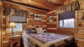 A cabin-style bedroom with a wooden interior, a bed with a quilt featuring animal prints, a nightstand with a lamp, and a window with blinds. - Cozy 2-bedroom cabin in beautiful Sevierville with fishing pond (Sevierville)