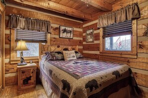 A cabin-style bedroom with a wooden interior, a bed with a quilt featuring animal prints, a nightstand with a lamp, and a window with blinds. - Cozy 2-bedroom cabin in beautiful Sevierville with fishing pond (Sevierville)