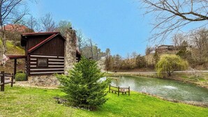 A rustic cabin with a stone foundation, a wooden deck, and a pond in the background. - Cozy 2-bedroom cabin in beautiful Sevierville with fishing pond (Sevierville)