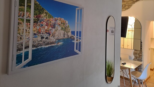 A room with a large window displaying a coastal town, a dining area with a table and chairs, and a mirror on the wall. - Manarola Sunset House | 2 Bathrooms | In the Heart of Cinque Terre (Manarola)