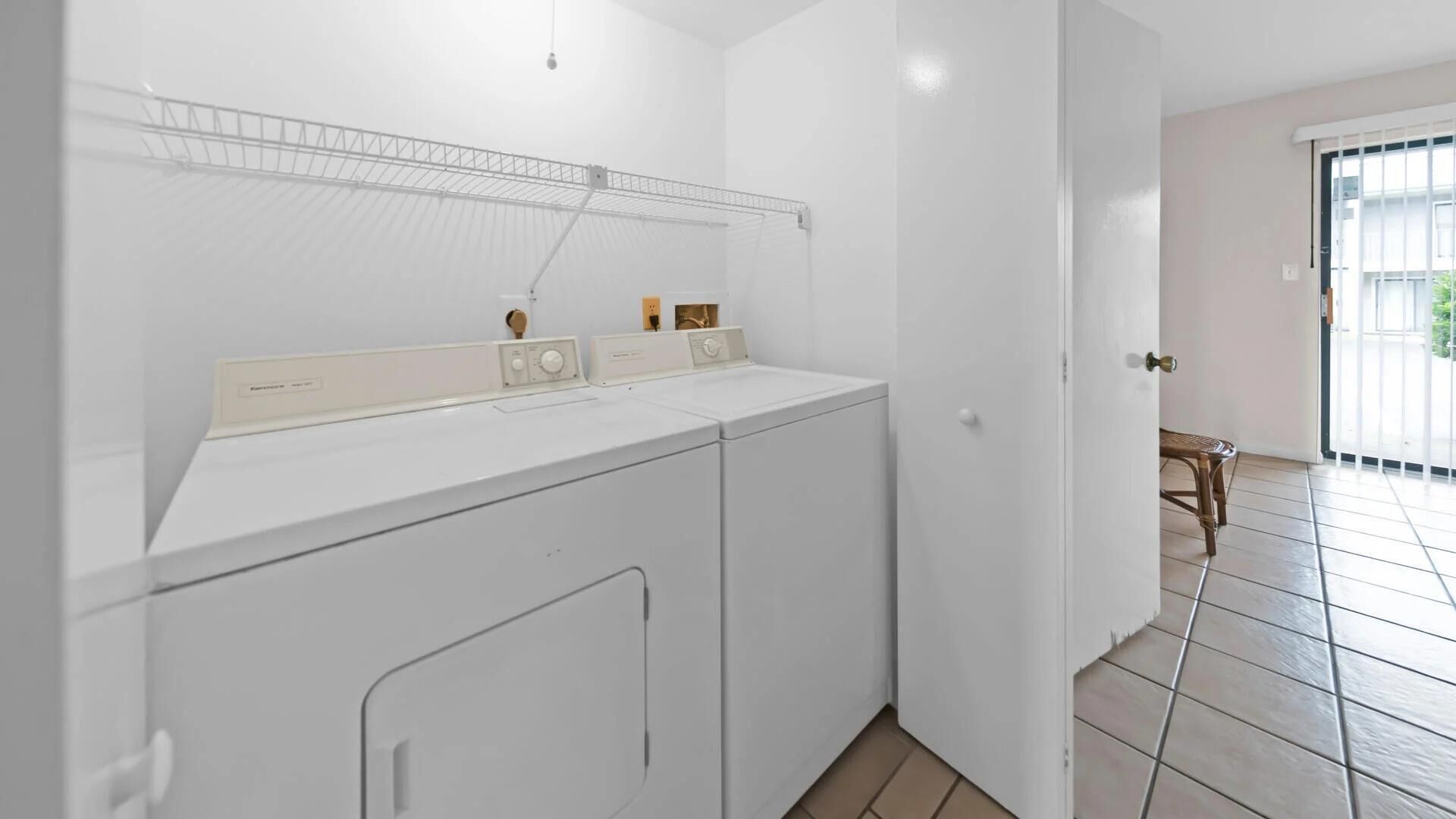 A laundry area with a washing machine and dryer, a metal shelf above the machines, and a door leading to a balcony.